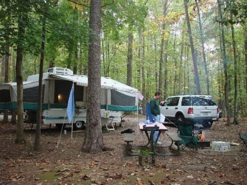 A campsite in a forest with a pop-up camper, a picnic table, and a person standing near a fire pit.