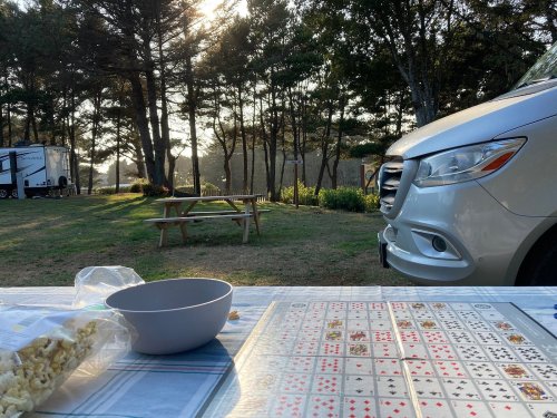 A picnic table in a campsite with a game board, popcorn, and a silver vehicle in the background under a sunny sky.