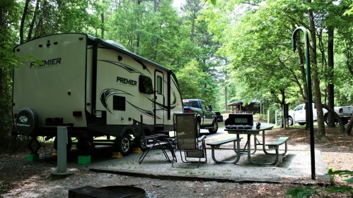 A camper parked in a wooded campsite with a picnic table, grill, and nearby vehicles under lush green trees.