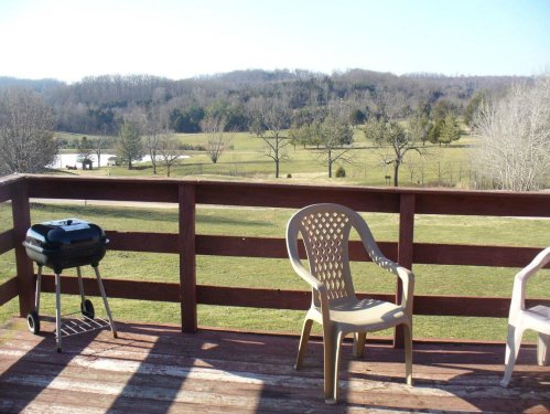 A wooden deck with two chairs and a small grill, overlooking a green landscape and distant hills under a clear sky.