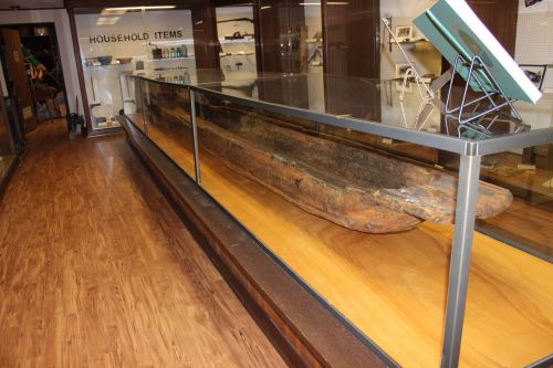 A long, weathered wooden artifact displayed in a glass case within a museum, with wooden flooring and shelves in the background.