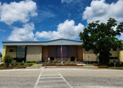A modern building with a blue roof, large windows, and a flagpole, set against a partly cloudy sky.