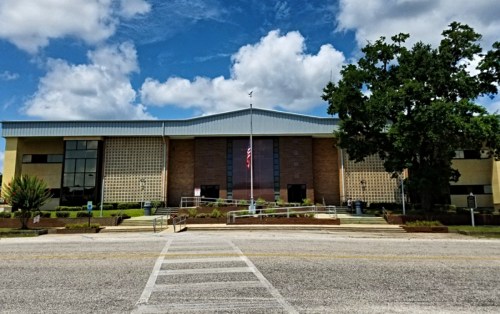 A modern building with a blue roof, large windows, and an American flag in front, set against a partly cloudy sky.