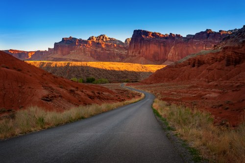 A winding road leads through red rock formations under a clear blue sky, with sunlight illuminating the landscape.