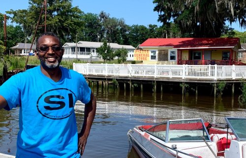 A smiling man in a blue shirt stands by a dock with a boat, overlooking a scenic waterfront and a colorful general store.