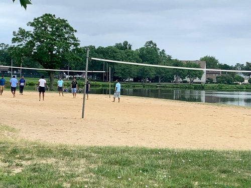 A sandy volleyball court by a lake, with several people playing and trees in the background under a cloudy sky.