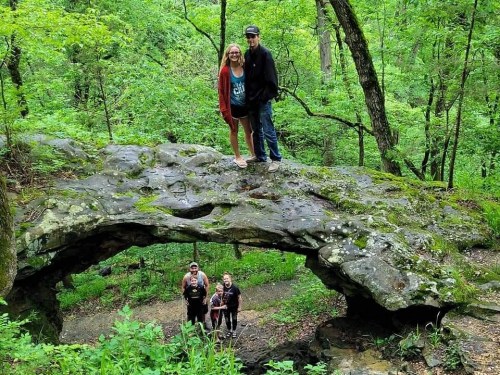 A couple stands on a moss-covered rock arch in a lush green forest, with two people below them on a trail.