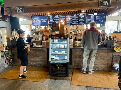 A coffee shop counter with a menu board, a customer ordering, and a display fridge with snacks.