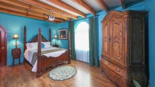 A cozy bedroom with a four-poster bed, wooden furniture, turquoise walls, and natural light from a large window.
