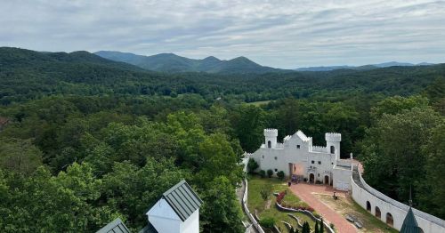 A scenic view of a white castle surrounded by lush green trees and mountains under a cloudy sky.