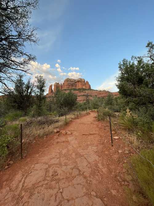 A rocky path leads through greenery towards towering red rock formations under a blue sky with scattered clouds.