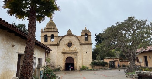 Historic mission building with a bell tower, surrounded by trees and a courtyard, under a cloudy sky.