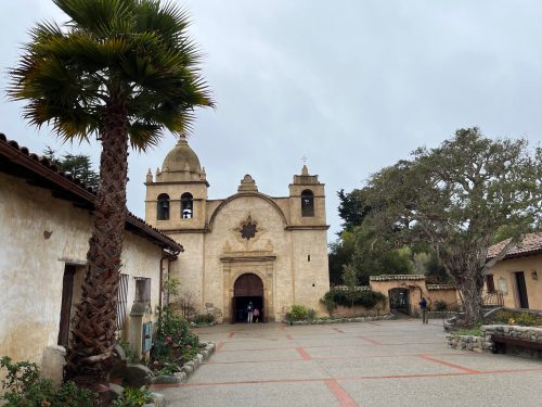 Historic mission building with a palm tree in the foreground, set in a courtyard under a cloudy sky.
