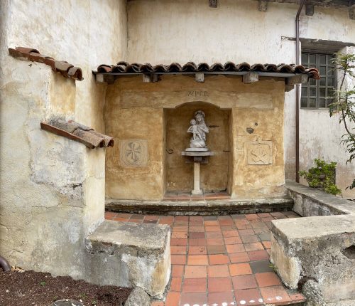A weathered stone alcove with a statue, surrounded by textured walls and a tiled floor, in a rustic outdoor setting.