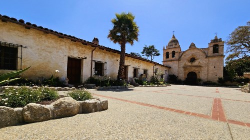 A historic mission building with a bell tower, surrounded by gardens and a clear blue sky.