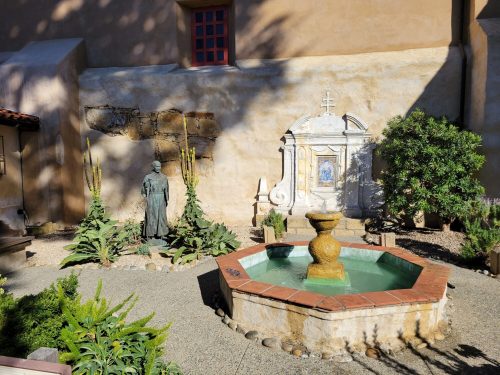 A serene courtyard featuring a fountain, stone wall, and greenery, with a statue in the background.