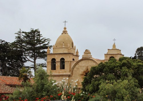 Historic building with domed roofs and crosses, surrounded by greenery and flowers under a cloudy sky.