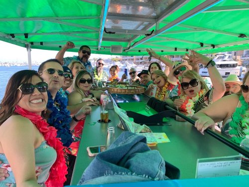 A lively group of people in tropical attire celebrating on a boat, enjoying drinks and smiling under a green canopy.
