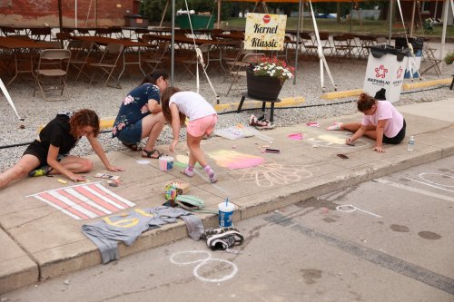 Four people, including children, are drawing with chalk on a sidewalk, surrounded by tables and a sign for a festival.