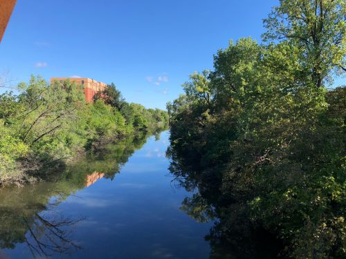 A calm river reflects trees and a building under a clear blue sky.