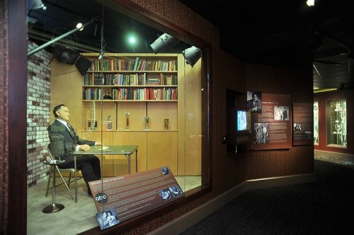A museum exhibit featuring a lifelike mannequin seated at a desk, surrounded by bookshelves and display panels.