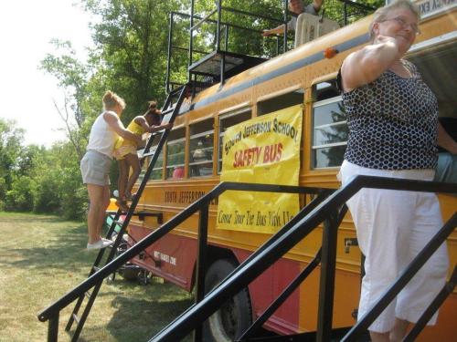 A yellow school bus with a safety banner, children climbing stairs, and adults supervising in a grassy area.