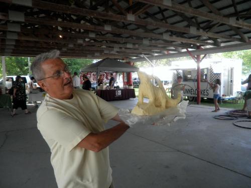 A man holds a large sculpture made of butter, smiling in a covered outdoor space with vendors in the background.