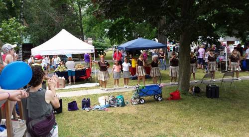 A group of performers in skirts stands on grass, with tents and a crowd in the background at an outdoor event.
