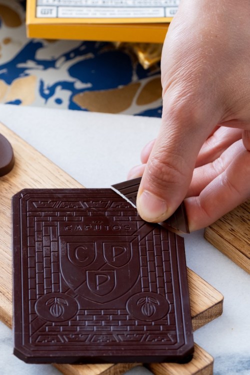A hand breaking a piece off a large chocolate bar on a wooden board, with other chocolate pieces in the background.