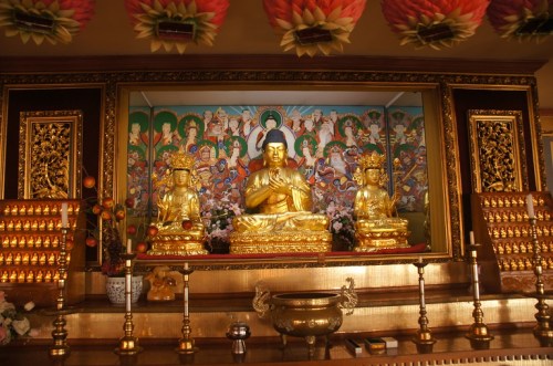 Golden Buddha statue surrounded by two smaller statues, with a colorful mural and offerings in a serene temple setting.