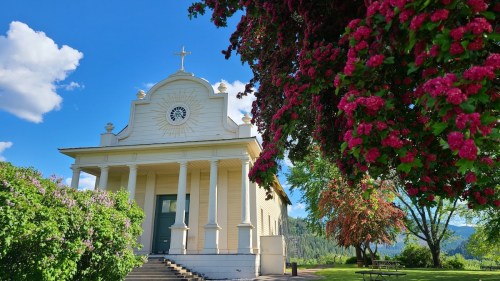 A white church with columns, a clock, and a cross, surrounded by vibrant flowers and lush greenery under a blue sky.