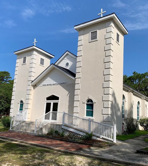 A white church building with two towers, a ramp entrance, and crosses on top, surrounded by greenery and blue sky.