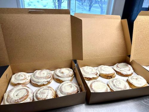 Two open boxes containing freshly baked cinnamon rolls with cream cheese frosting, set against a snowy window backdrop.