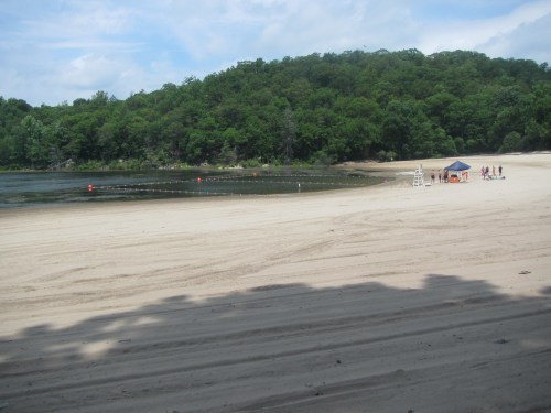 A sandy beach with a lifeguard stand, surrounded by green hills and a calm water area, under a partly cloudy sky.