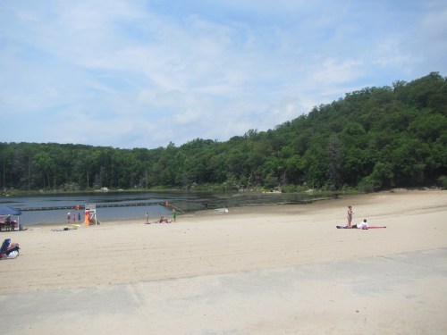 A sandy beach with a calm lake, surrounded by green trees and a wooden dock in the background. People relax on the shore.