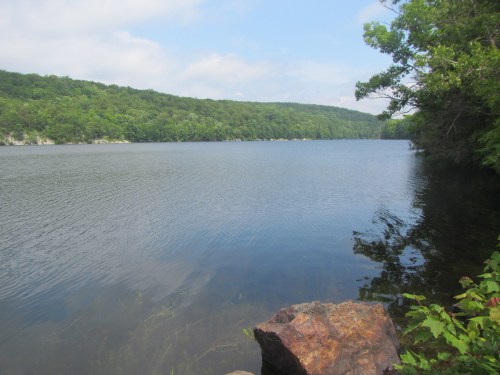 A serene lake surrounded by lush green hills under a partly cloudy sky, with a rocky shore in the foreground.