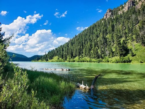 A serene lake surrounded by lush green hills and trees, with a clear blue sky and fluffy white clouds above.