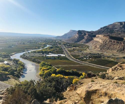 A scenic view of a valley with a river, fields, and mountains under a clear blue sky.