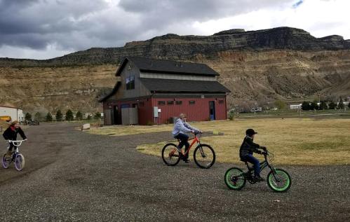 Three children ride bicycles on a gravel path near a red barn and rocky hills under a cloudy sky.