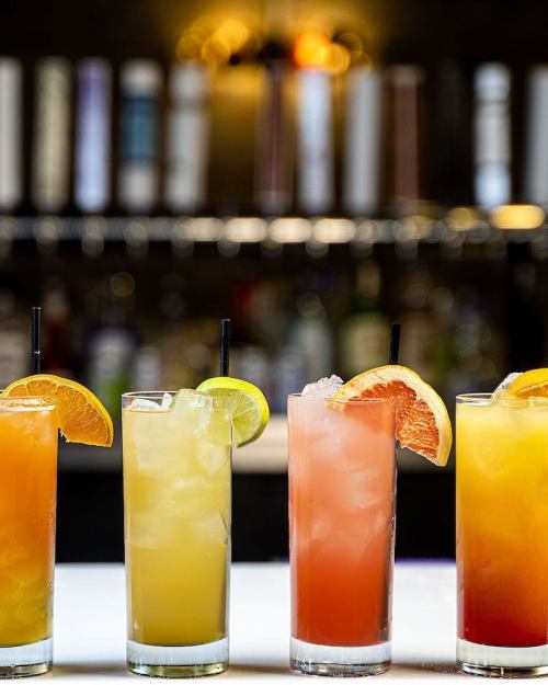 Four colorful cocktails with fruit garnishes, displayed on a bar counter with blurred bottles in the background.