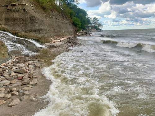 Waves gently crash on a rocky shoreline under a cloudy sky, with trees lining the cliff in the background.