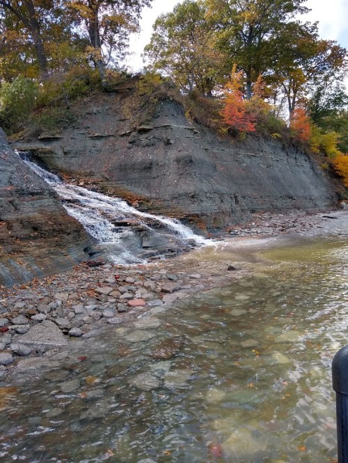 A small waterfall cascades down a rocky cliff into a calm body of water, surrounded by autumn foliage.