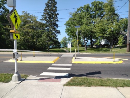 A crosswalk with yellow markings, surrounded by trees and a clear blue sky, near a road and pedestrian signs.