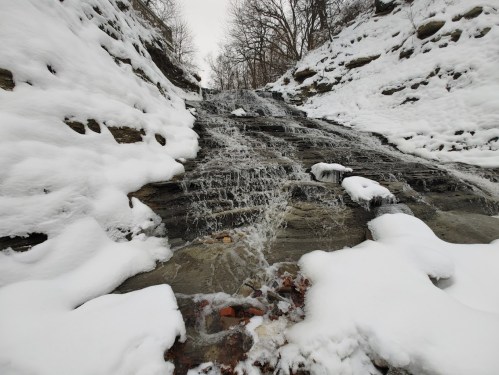 A snowy landscape featuring a small waterfall cascading down rocky terrain, surrounded by snow-covered ground.