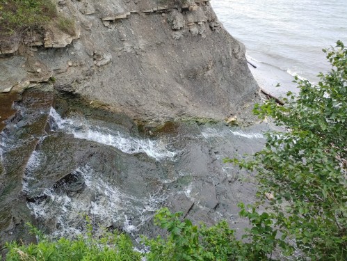 A rocky cliffside with cascading water flowing into a river, surrounded by greenery and a calm water surface.