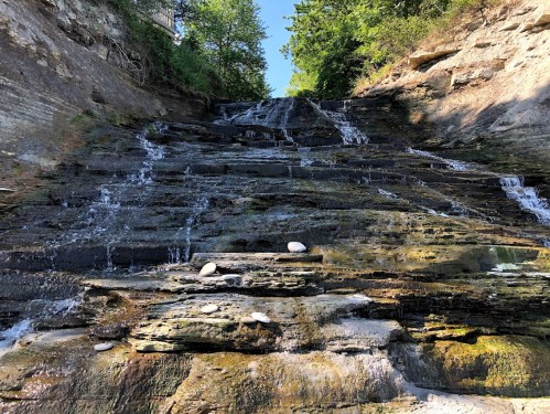 A serene waterfall cascading down rocky layers, surrounded by lush greenery under a clear blue sky.