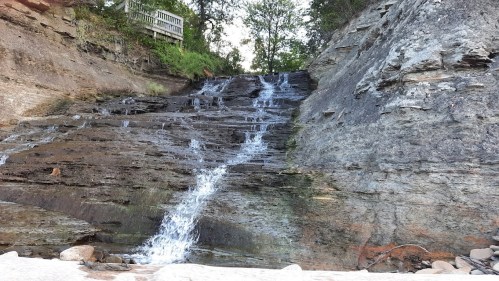 A cascading waterfall flows down a rocky cliff, surrounded by greenery and a wooden railing above.