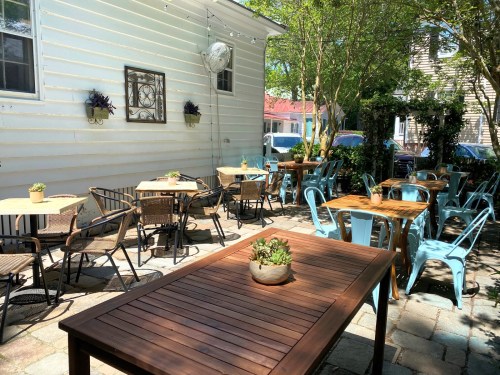 Outdoor dining area with wooden and metal tables, surrounded by greenery and a white building in the background.