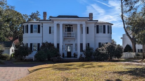 A large, white, two-story house with black shutters and a front porch, surrounded by greenery and a clear blue sky.