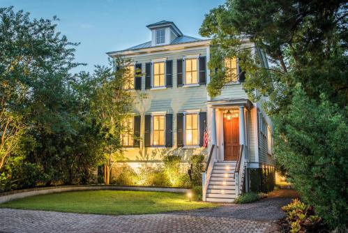 A charming two-story house with a porch, surrounded by greenery and illuminated at dusk. An American flag hangs by the door.